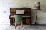 A vintage wooden upright piano stands against a textured, off-white wall. An open music sheet rests on the piano's stand. In front of the piano is a wooden chair with a green upholstered seat. A tall lamp with a unique shade patterned with nature scenes is positioned to the right of the piano, standing on an ornate, twisted stand.