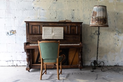 A vintage wooden upright piano stands against a textured, off-white wall. An open music sheet rests on the piano's stand. In front of the piano is a wooden chair with a green upholstered seat. A tall lamp with a unique shade patterned with nature scenes is positioned to the right of the piano, standing on an ornate, twisted stand.