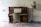 A vintage wooden upright piano stands against a textured, off-white wall. An open music sheet rests on the piano's stand. In front of the piano is a wooden chair with a green upholstered seat. A tall lamp with a unique shade patterned with nature scenes is positioned to the right of the piano, standing on an ornate, twisted stand.