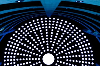 A circular array of glowing white dots set against a dark background, forming a mesmerizing pattern. The structure appears to be inside a tunnel or cylindrical object with a symmetrical design, filled with evenly spaced holes that allow light to pass through.