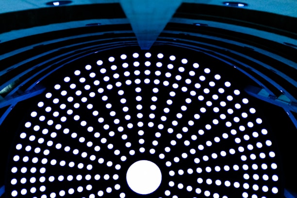 A circular array of glowing white dots set against a dark background, forming a mesmerizing pattern. The structure appears to be inside a tunnel or cylindrical object with a symmetrical design, filled with evenly spaced holes that allow light to pass through.