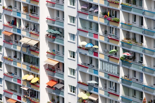 A multi-story residential building facade with numerous balconies adorned with colorful awnings and vibrant flower boxes. Some balconies feature umbrellas and potted plants, adding to the lively atmosphere. The building is characterized by a modern architectural design with a repetitive pattern of windows and railing bars.
