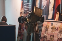 An exhibition setting with a vintage-looking telescope or similar optical instrument displayed prominently in the foreground. Posters and informational banners are visible in the background, featuring images and text related to educational or professional topics. A person is partially visible, standing near a partition.