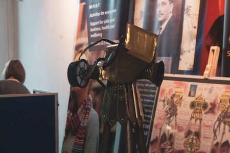 An exhibition setting with a vintage-looking telescope or similar optical instrument displayed prominently in the foreground. Posters and informational banners are visible in the background, featuring images and text related to educational or professional topics. A person is partially visible, standing near a partition.
