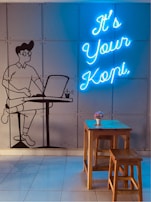 A wall features a mural of a person sitting at a table using a laptop, next to them is a cup. The words 'It's Your Kopi' are displayed in bright blue neon lights. In front of the wall, there is a wooden table with two stools and a small flower arrangement in a white pot.