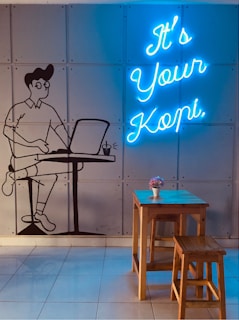 A wall features a mural of a person sitting at a table using a laptop, next to them is a cup. The words 'It's Your Kopi' are displayed in bright blue neon lights. In front of the wall, there is a wooden table with two stools and a small flower arrangement in a white pot.