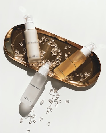 Three skincare bottles are arranged on a gold tray surrounded by small, clear crystal-like stones. The bottles have minimalistic branding with simple fonts, enhancing the luxurious aesthetic. The tray casts a soft shadow, indicating bright, direct lighting.