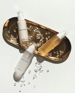 Three skincare bottles are arranged on a gold tray surrounded by small, clear crystal-like stones. The bottles have minimalistic branding with simple fonts, enhancing the luxurious aesthetic. The tray casts a soft shadow, indicating bright, direct lighting.