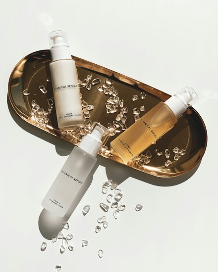 Three skincare bottles are arranged on a gold tray surrounded by small, clear crystal-like stones. The bottles have minimalistic branding with simple fonts, enhancing the luxurious aesthetic. The tray casts a soft shadow, indicating bright, direct lighting.