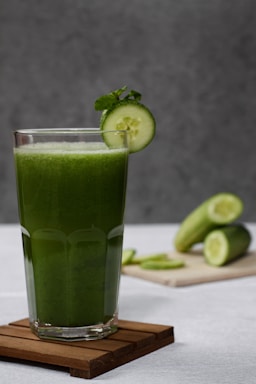 A tall glass filled with a vibrant green smoothie, garnished with a slice of cucumber and mint leaf. The glass is placed on a wooden coaster, with more cucumber slices and whole cucumbers in the background on a wooden board. The setting has a neutral, soft gray background.
