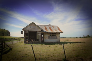 An old, weathered house stands isolated in a grassy field under a clear blue sky. The building appears neglected, with rusting metal roofing and peeling paint. A sign on the wall reads 'HOUSE FOR RENT'. Surrounding the house is a wooden fence, and a dirt path leads away into the distance beside a crop field.