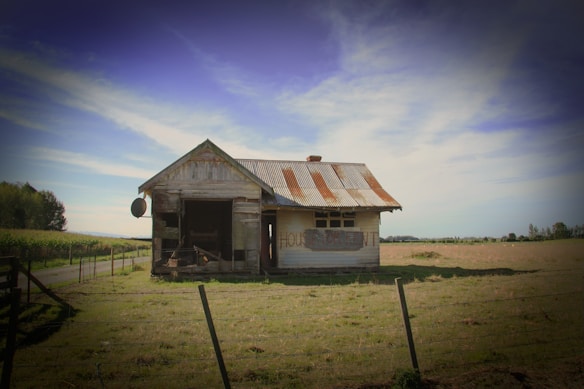 An old, weathered house stands isolated in a grassy field under a clear blue sky. The building appears neglected, with rusting metal roofing and peeling paint. A sign on the wall reads 'HOUSE FOR RENT'. Surrounding the house is a wooden fence, and a dirt path leads away into the distance beside a crop field.