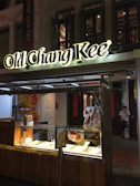 A small street food kiosk with a prominent 'Old Chang Kee' sign illuminated at night. The food display case is below the sign, with various food items and promotional posters on display. A person is seated in the background near the doorway.