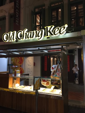 A small street food kiosk with a prominent 'Old Chang Kee' sign illuminated at night. The food display case is below the sign, with various food items and promotional posters on display. A person is seated in the background near the doorway.