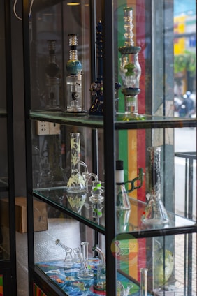 A display case filled with various glass bongs and smoking apparatuses arranged on multiple shelves. The items are intricately designed and some have colorful patterns. The background shows a street scene outside with blurred people and buildings.