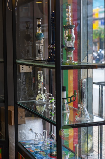 A display case filled with various glass bongs and smoking apparatuses arranged on multiple shelves. The items are intricately designed and some have colorful patterns. The background shows a street scene outside with blurred people and buildings.