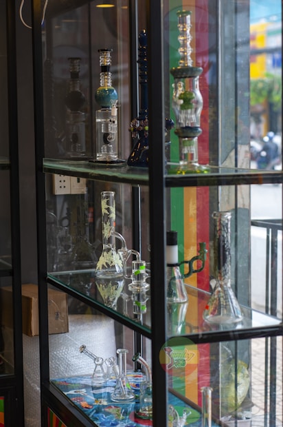 A display case filled with various glass bongs and smoking apparatuses arranged on multiple shelves. The items are intricately designed and some have colorful patterns. The background shows a street scene outside with blurred people and buildings.