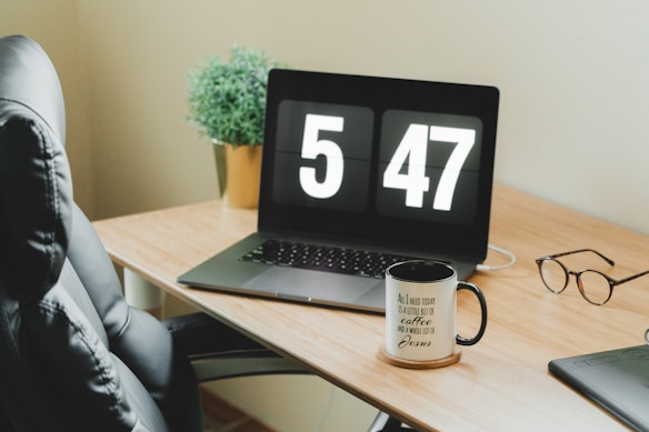 A modern workspace features a wooden desk with a black swivel chair. On the desk, a laptop displays the time '5:47' in large digital numbers. Nearby, a ceramic mug with motivational text sits on a coaster, and a pair of glasses rests beside a potted plant. The scene exudes a tidy and minimalist aesthetic.
