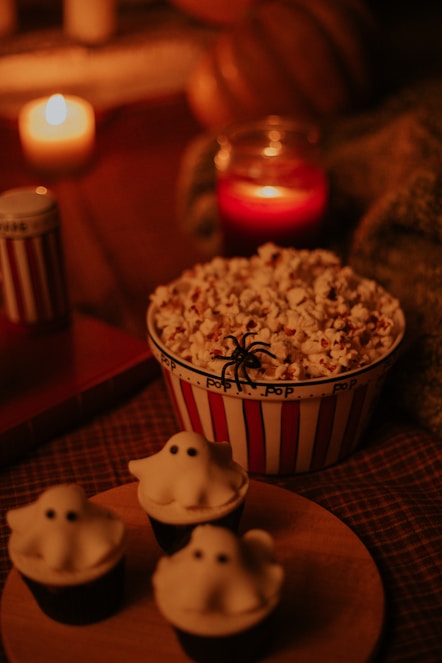 A cozy and warm scene featuring a striped popcorn bucket topped with a plastic spider, surrounded by candles and blankets. Three ghost-themed cupcakes are placed on a wooden platter, enhancing the Halloween theme.