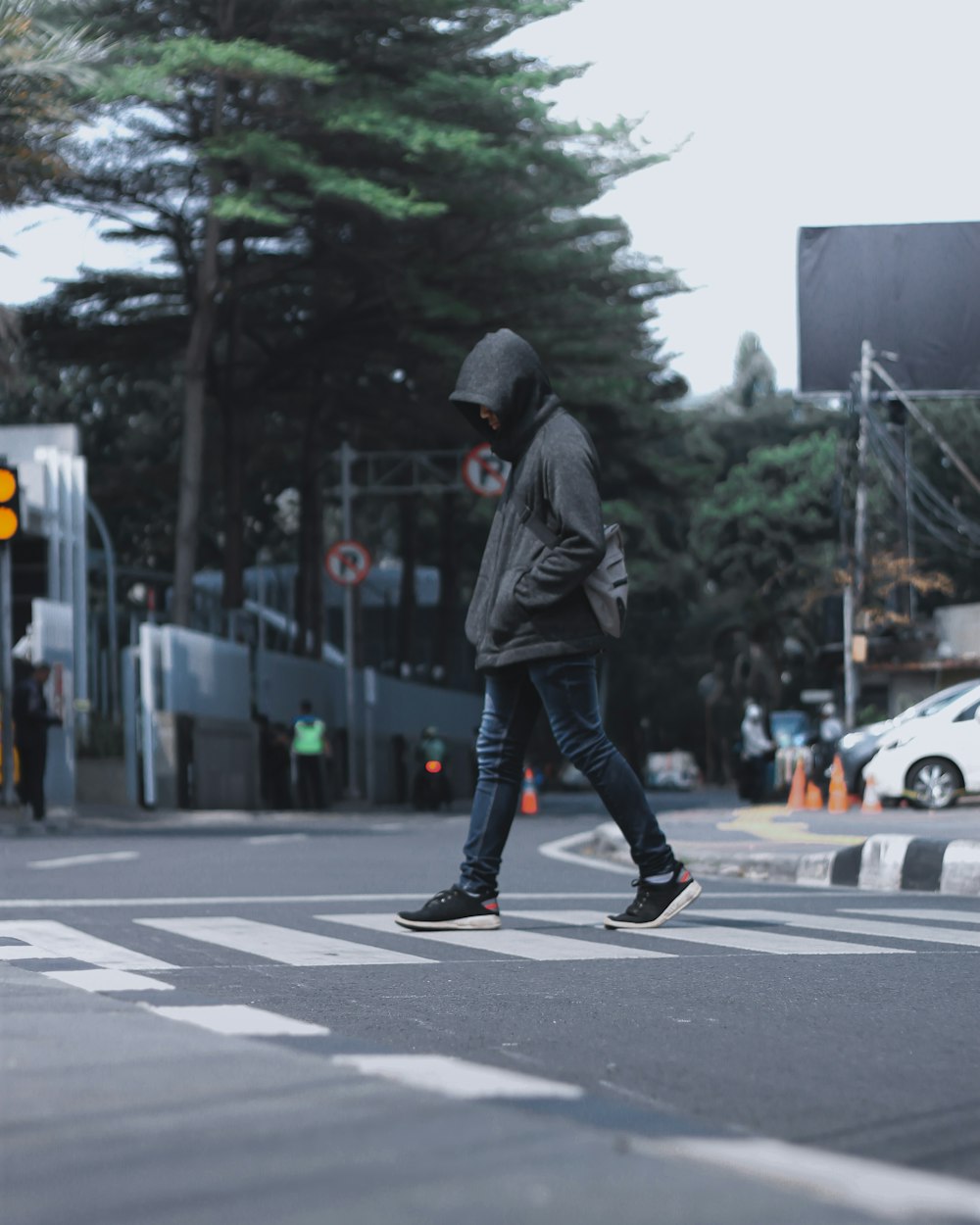 photography of man wearing black hooded jacket and blue denim jeans passing by the pedestrian lane