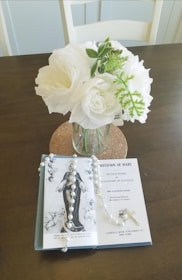 A vase with white flowers and green leaves is placed on a table. Nearby is an open book titled 'Imitation of Mary' along with a pearl rosary adorned with a cross. The setting is calm and serene, with soft lighting enhancing the peaceful atmosphere.