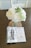 A vase with white flowers and green leaves is placed on a table. Nearby is an open book titled 'Imitation of Mary' along with a pearl rosary adorned with a cross. The setting is calm and serene, with soft lighting enhancing the peaceful atmosphere.