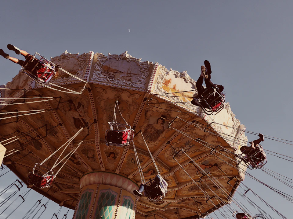 The image depicts a large, ornate carousel with a white and gold color scheme, featuring intricate designs on its sides and top. The carousel is filled with people enjoying their ride, some seated while others stand holding onto the ropes for balance. The sky above them is clear blue, providing a beautiful backdrop to this lively scene.