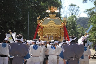 A group of people dressed in traditional attire are carrying an ornate golden portable shrine through a wooded area. The shrine is adorned with red tassels and intricate designs, while the individuals wear robes with blue insignias and headbands. Sunlight filters through the trees, creating a serene and festive scene.