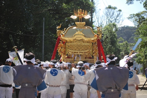 A group of people dressed in traditional attire are carrying an ornate golden portable shrine through a wooded area. The shrine is adorned with red tassels and intricate designs, while the individuals wear robes with blue insignias and headbands. Sunlight filters through the trees, creating a serene and festive scene.