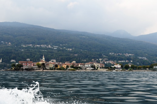 A serene landscape featuring a small town nestled at the base of rolling hills and mountains. The town's buildings, with a prominent church steeple, are bordered by lush greenery. The foreground shows a body of water with gentle waves, and there is overcast weather, resulting in a calm and muted atmosphere.