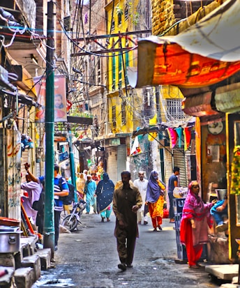 A bustling narrow street with people walking in various colorful traditional attire. The street is lined with small shops and vibrant banners. Buildings show vibrant shades of yellow and brown, and there are numerous electrical wires crisscrossing overhead.