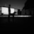 A silhouette of a person and a dog standing in front of an outdoor cinema setup. The image is predominantly in black and white, highlighting the dark figures against a backdrop of chairs and an architectural building. The scene appears to be set during dusk or dawn, adding dramatic effects to the shadow play.