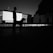 A silhouette of a person and a dog standing in front of an outdoor cinema setup. The image is predominantly in black and white, highlighting the dark figures against a backdrop of chairs and an architectural building. The scene appears to be set during dusk or dawn, adding dramatic effects to the shadow play.