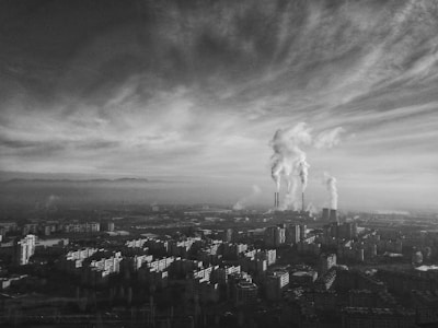 A black and white image of a cityscape with industrial buildings. Smoke billows from several tall chimneys, indicating air pollution. The sky is filled with clouds that seem to blend with the smoke.