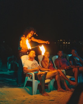 A performer is engaged in a fire show, holding torches with flames in front of an audience sitting on the beach in plastic chairs. The atmosphere is lively and the sand is illuminated by the orange glow of the fire. The audience consists of casually dressed people who appear relaxed and entertained.