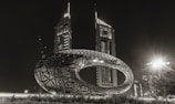 A futuristic and uniquely shaped torus structure with intricate Arabic calligraphy is prominently displayed in front of two modern, tall skyscrapers. The scene is illuminated at night, highlighting the architecture and artistic design against the dark sky.