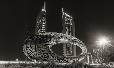 A futuristic and uniquely shaped torus structure with intricate Arabic calligraphy is prominently displayed in front of two modern, tall skyscrapers. The scene is illuminated at night, highlighting the architecture and artistic design against the dark sky.