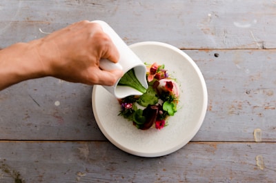 A hand is pouring green sauce from a white jug onto an artfully arranged salad with various colorful garnishes on a white plate. The setting is a rustic wooden table that adds a natural feel to the presentation.