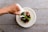 A hand is pouring green sauce from a white jug onto an artfully arranged salad with various colorful garnishes on a white plate. The setting is a rustic wooden table that adds a natural feel to the presentation.