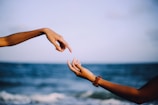 Two hands are positioned towards each other against a blurred background of the ocean and sky, suggesting connection or reaching out.