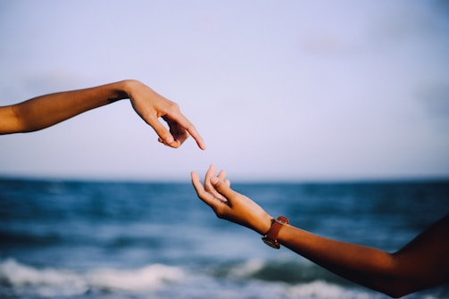 Two hands are positioned towards each other against a blurred background of the ocean and sky, suggesting connection or reaching out.