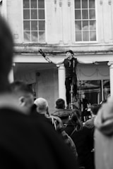 A street performer is balancing on a unicycle, holding juggling clubs in each hand, with a small crowd gathered around watching. The setting appears to be an outdoor area with large windows and columns in the background.
