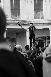 A street performer is balancing on a unicycle, holding juggling clubs in each hand, with a small crowd gathered around watching. The setting appears to be an outdoor area with large windows and columns in the background.