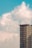 A tall residential building with multiple windows set against a backdrop of a bright, partly cloudy sky. The building is modern with numerous floors and balconies, situated on the right side of the image.