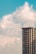 A tall residential building with multiple windows set against a backdrop of a bright, partly cloudy sky. The building is modern with numerous floors and balconies, situated on the right side of the image.