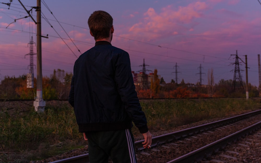 man wearing black jacket standing near train railway under blue and orange sky during daytime