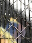 A pattern of spray-painted metal bars behind a chain-link fence. The bars feature a gradient of colors, including black, white, yellow, and blue, with a speckled texture. The chain-link fence is intertwined with the metal bars, creating a layered, abstract composition.