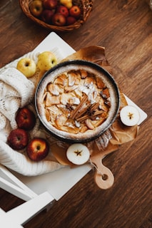 A rustic apple pie garnished with cinnamon sticks is placed on a cutting board, surrounded by whole red and yellow apples. The setup rests on a white chair with a soft knitted blanket, creating a cozy autumnal scene. In the background, a basket filled with various apples is visible on a wooden floor.