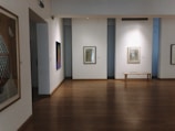 A quiet and empty art gallery room featuring wooden floors and white walls. Several framed artworks are displayed on the walls, with one colorful painting on the left and another black and white piece directly ahead next to a simple wooden bench.