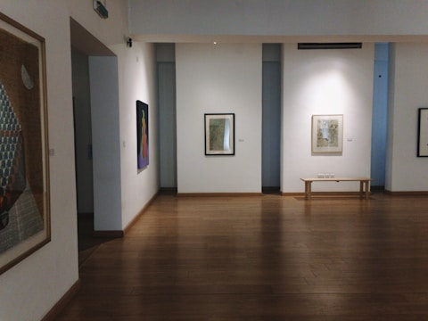 A quiet and empty art gallery room featuring wooden floors and white walls. Several framed artworks are displayed on the walls, with one colorful painting on the left and another black and white piece directly ahead next to a simple wooden bench.